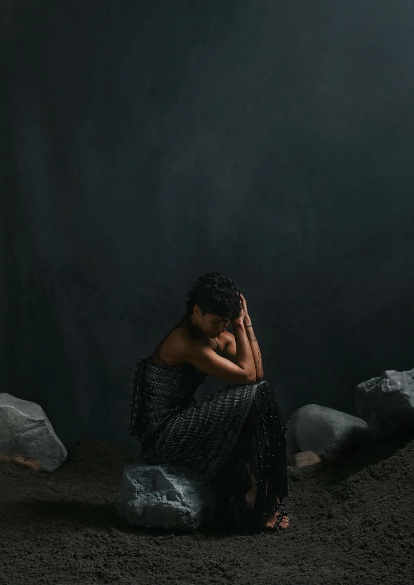 Maria Anouk seated on a rock in a dark setting, wearing a textured black garment from Binhe. Design assistance and product design by Storm Nijhuis for Zyanya Keizer.