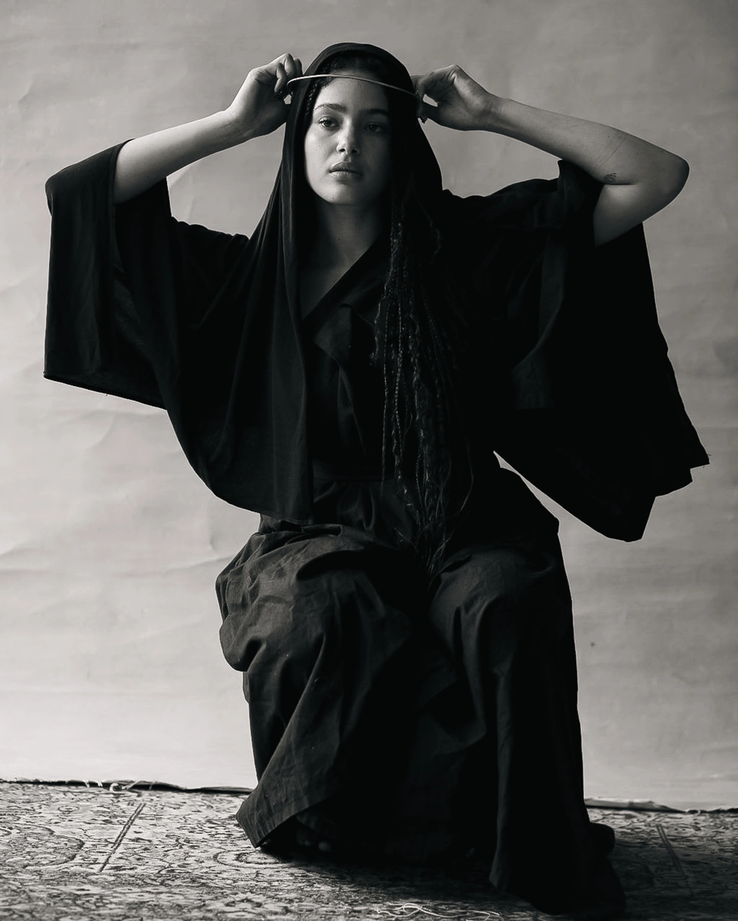 Louise Aniba seated in a black draped garment, holding a circular headpiece above her head. Styling by Storm Nijhuis for Divine Feminine.