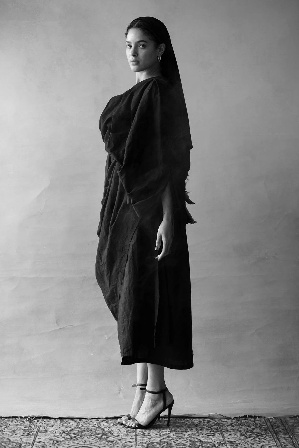 Louise Aniba standing in a sculptural black dress, with her heels visible and smoke around the garment. Styling by Storm Nijhuis for Divine Feminine.